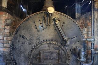 Façade rivetée d'une ancienne chaudière à vapeur avec manomètre et niveau d'eau, dans une salle en briques
